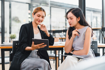 Two Asian women, one in black blazer and other in sleeveless top, sit at table in modern office, discussing work while looking at digital tablet, appearing focused and collaborative