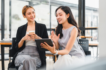 Two Asian women, one holding smartphone and other using digital tablet, sit together in modern office, smiling and discussing work in collaborative, friendly atmosphere