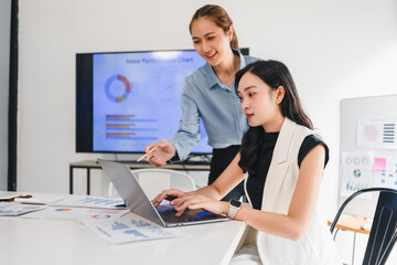 Two Asian businesswomen working together in modern office, discussing data analysis with charts and...