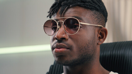 Portrait African American guy man ethnic male putting on sunglasses glasses wearing looking at camera sitting in futuristic white studio futurism innovation stylish fashion posing indoors sit in chair