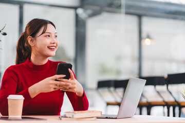 Young woman red sweater sitting at desk holding smartphone smiling working on laptop coffee cup books modern office natural light cheerful productivity