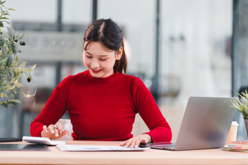 Young Asian woman in red sweater working at desk with laptop, tablet, and documents, smiling and focused in modern office environment, showing productivity and positive energy