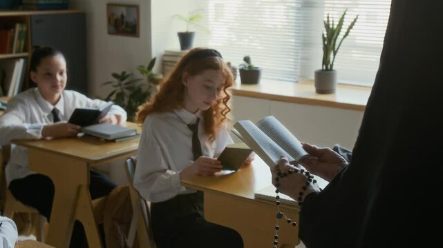 Slowmo of multiethnic diligent teen girls in school uniform sitting at single desks listening carefully to unrecognizable nun in veil reading Bible out loud during catechism class in Catholic school