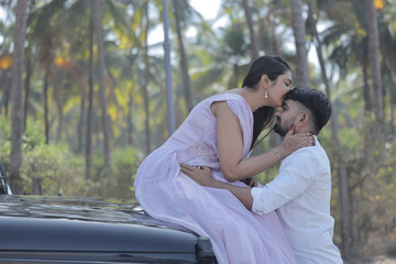 Naklejka premium A cheerful Indian couple shares a romantic moment on a palm-lined road, standing in front of a black SUV. The woman is wearing a flowing lavender gown, and the man embraces her from behind with a joyf