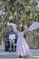 Obraz premium A cheerful Indian couple shares a romantic moment on a palm-lined road, standing in front of a black SUV. The woman is wearing a flowing lavender gown, and the man embraces her from behind with a joyf
