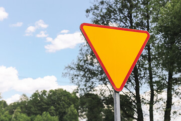 Bright yellow traffic sign. Triangular road warning symbol. Urban infrastructure with trees. Give way triangle sign. Safety signal in countryside. Driving rules and signage visibility reminder.