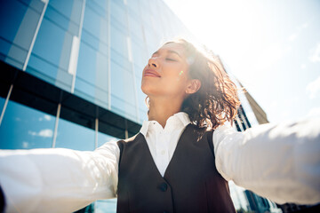 Confident young businesswoman enjoys a sunny day outdoors near a modern office building, embodying...