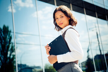 Beautiful confident businesswoman with clipboard standing outdoors near modern office building on a sunny day