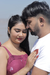 A cheerful Indian couple enjoys a sunny day at the beach, holding hands while walking along the shoreline. The woman is dressed in a casual maroon dress, and the man wears a white t-shirt with grey sh