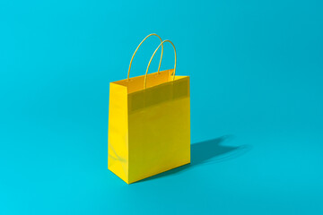 Shopping paper bag standing on the turquoise blue background with copy space. Minimalist photo of yellow bag with harsh shadow central composition. Summer sale and clearance sale concepts.