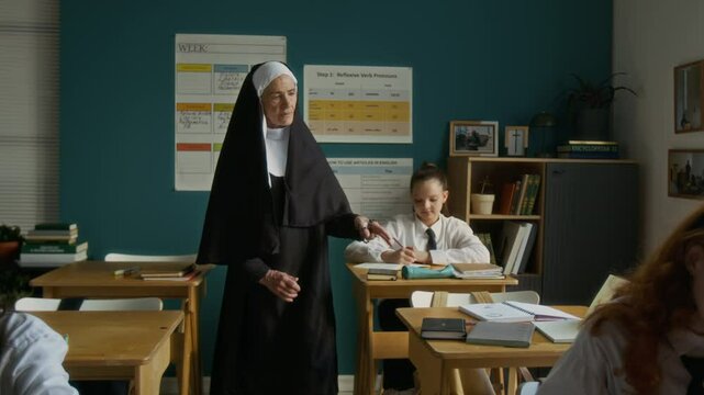 Slowmo shot of senior Caucasian nun teacher in veil supervising work of multiethnic teen student girls studying in modern well-equipped classroom in religious school