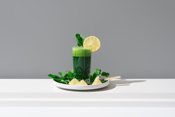 Jugo de espinacas adornado con una rodaja de limón y hoja de espinaca sobre una mesa blanca y fondo gris	