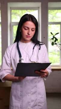 Medical professional checks clipboard, looks up and nods at camera, then refocuses on papers. Concept of female healthcare professionals, medical office workflow