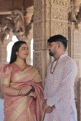 An Indian couple poses gracefully at a beautifully carved Hindu temple. The woman is wearing a rich...