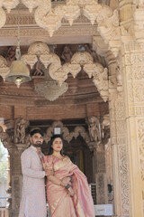 An Indian couple poses gracefully at a beautifully carved Hindu temple. The woman is wearing a rich golden-pink silk saree adorned with jewelry and bangles, while the man is dressed in an elegant ethn