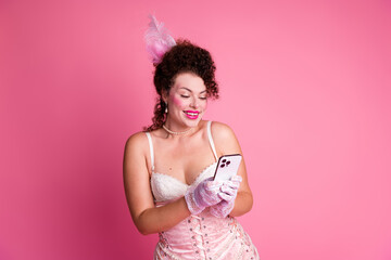 Young woman in elegant baroque costume with pink feathers holding a smartphone against a vibrant...