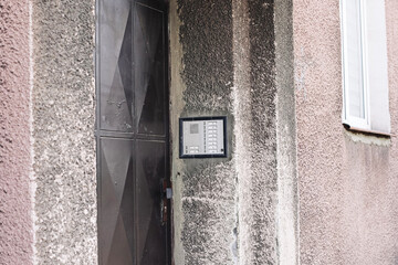 Old apartment intercom system. Entry phone with buttons on weathered wall. Vintage access control panel near metal door. Residential building entrance with textured concrete and worn facade.