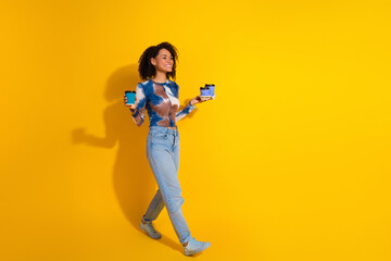 Stylish young woman holding coffee and dessert walking confidently against vibrant yellow background