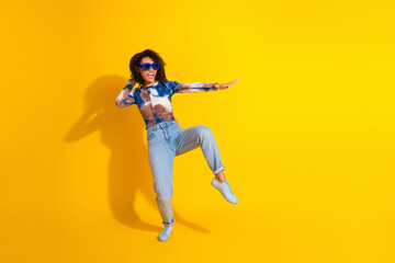 Young woman joyfully dancing against a vibrant yellow background wearing sunglasses and casual...
