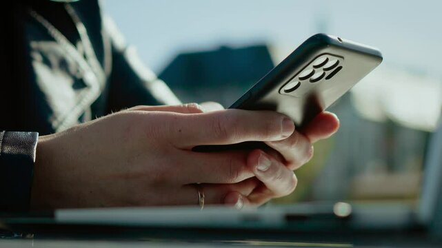 Female worker typing on mobile phone outdoors. Woman using smartphone near laptop in daylight. Remote employee sending message on digital device. Unrecognizable user interacting with screen in open