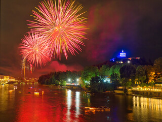 Fireworks over the Po River in Turin, Italy, during the San Giovanni celebration, with illuminated boats, Monte dei Cappuccini, and the Church of Santa Maria al Monte lit in blue.