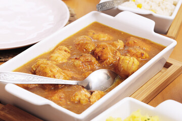 Traditional meat dish in sauce. Tasty homemade meal in white ceramic tray. Comfort food with rich gravy. Warm lunch served with rice. Rustic kitchen table setup with spoon and natural lighting.