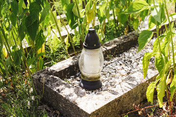 Glass candle on old grave. Cemetery lantern without flame among green plants. Peaceful burial place in summer light. Symbol of memory and silence. Nature surrounding forgotten resting spot.