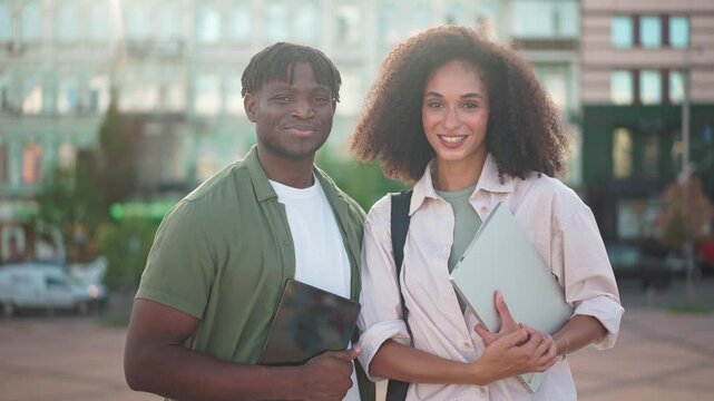 Smiling people posing in front of camera. Urban sunny background. African American man holding tablet devise while standing next to beautiful curly woman with laptop in her hands. Technology concept.