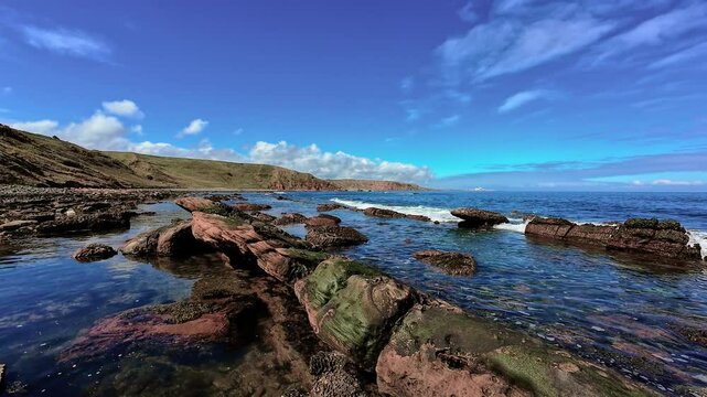 Waves splashing over red and green rocks along clear sea. Coastal tide rolling onto weathered stones beneath blue sky. Surf breaking gently on rocky shoreline near hills. Ocean meeting sculpted rock