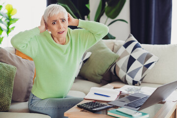 Mature woman at home experiencing stress while managing finances with a laptop and documents in a...