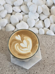 beautifully crafted latte with intricate foam art sits on marble surface, surrounded by smooth white pebbles, creating serene and inviting atmosphere