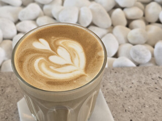 beautifully crafted latte with intricate foam art sits atop smooth surface, surrounded by white pebbles, creating serene and inviting atmosphere