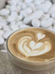 beautifully crafted latte with intricate foam art sits in glass cup, surrounded by backdrop of smooth white stones, creating serene and inviting atmosphere