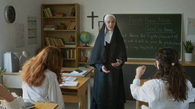 Motivated teen Caucasian student girl raising hand during class in religious school and reading Bible at blackboard in front of senior nun teacher and classmates