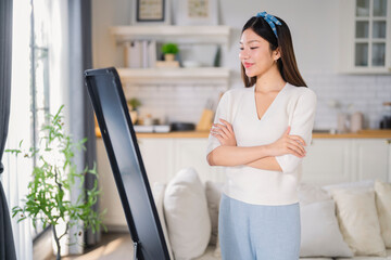 A confident, cheerful Asian woman smiles warmly as she looks at herself in the mirror at home,...