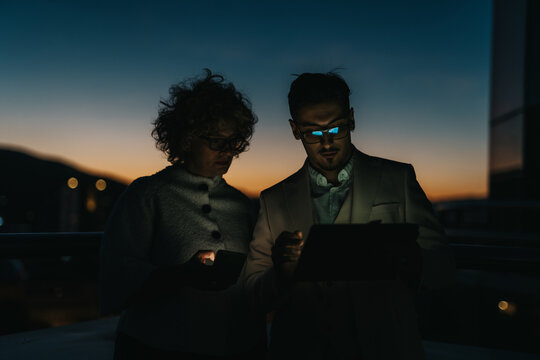 Two business people work together at night, analyzing data on a digital tablet. The background shows a serene sunset, highlighting the focused ambiance of their collaborative effort.