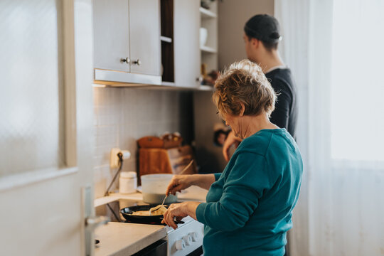 An elderly woman prepares a meal in a cozy kitchen, accompanied by a young man in the background.