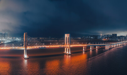 Aerial view of the illuminated bridge and cityscape over the reflective water at night in Macau.