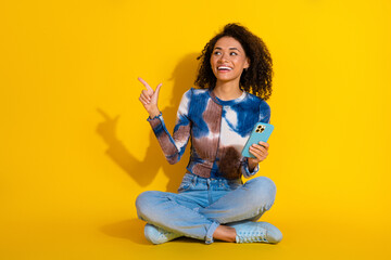 Young woman with curly hair holding a smartphone and pointing with a cheerful expression in a...