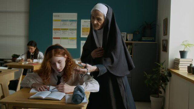 Elderly Caucasian nun in veil working as teacher in religious school and checking diverse student girls assignment during lesson in modern well-equipped classroom