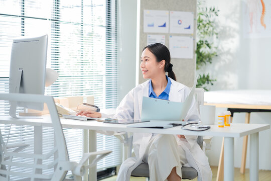 Portrait of a confident Asian female doctor smiling while working at her desk in a clinic, typing on her laptop and reviewing medical reports. Modern healthcare professional indoors with document file