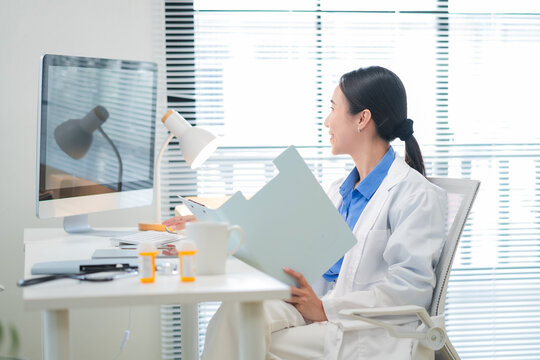 Portrait of a confident Asian female doctor smiling while working at her desk in a clinic, typing on her laptop and reviewing medical reports. Modern healthcare professional indoors with document file