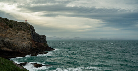 A lone lighthouse perched on a rugged cliff overlooks the choppy sea under a dramatic, overcast...
