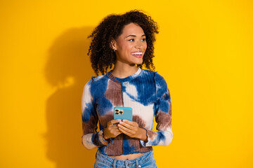 Cheerful young woman holding a phone, standing against a vibrant yellow background, showcasing...