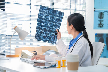 Asian female doctor or nurse in a white lab coat analyzing an X-ray scan in a medical clinic. She examines patient radiographs carefully, studying brain or lung images on film or screen, representing 