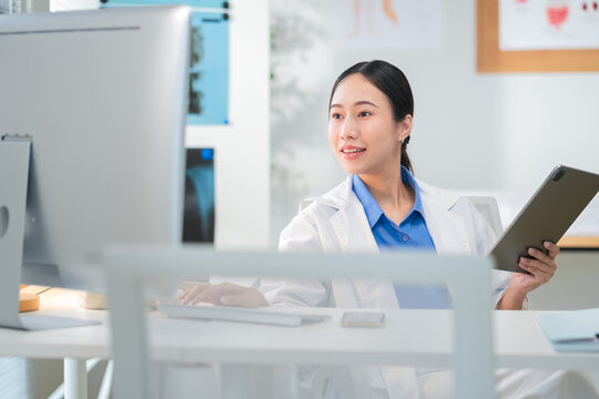 Portrait of a confident Asian female doctor smiling while working at her desk in a clinic, typing on her laptop and reviewing medical reports. Modern healthcare professional indoors with document file