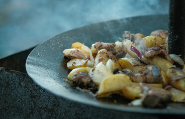 Savory outdoor-cooked meal of fried potatoes, bacon, and onions, sizzling on a black metal disc.