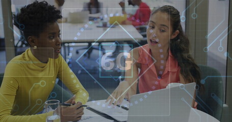 Collaborating two women discussing documents on laptop in office meeting room, with 6G overlay