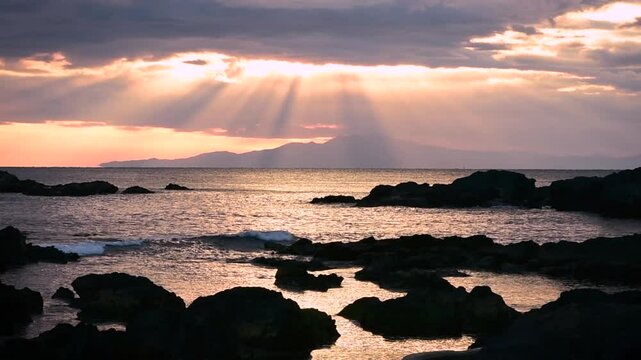 神奈川県三浦市の磯から望む夕日と光芒。岩場と海面に差し込む天使の梯子が幻想的な雰囲気を醸し出す風景。夕暮れ時の静けさと自然美を感じられる絶景の海岸線の映像。

