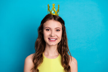 Cheerful brunette woman wearing a yellow dress with a charming crown on a vibrant blue background...
